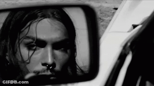 A Black And White Photo Of A Man With Long Hair And A Nose Ring Looking At Himself In A Rear View Mirror . GIF