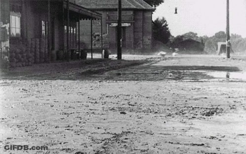 A Black And White Photo Of A Muddy Street In Front Of A Building That Says ' Warehouse ' On The Front GIF