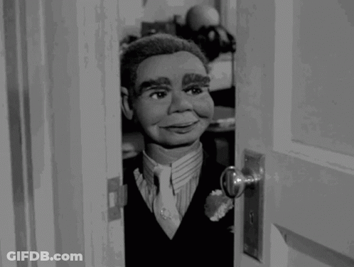 A Black And White Photo Of A Puppet In A Suit And Tie Looking Out Of A Door GIF