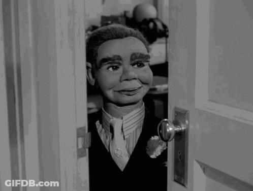 A Black And White Photo Of A Puppet Looking Out Of A Door GIF