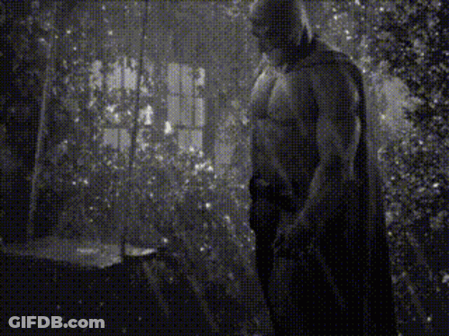 A Black And White Photo Of A Shirtless Man In A Cape Standing In A Room . GIF