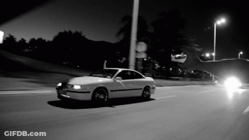 A Black And White Photo Of A White Car Driving Down A Street GIF