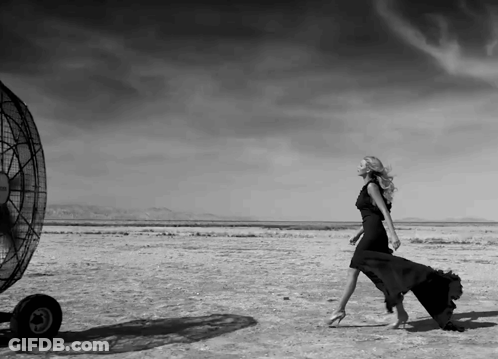 A Black And White Photo Of A Woman In A Black Dress Walking In The Desert GIF