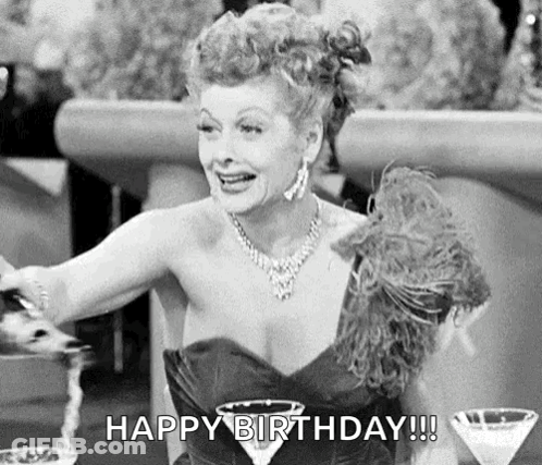 A Black And White Photo Of A Woman In A Dress Sitting At A Table With A Martini Glass And A Feather Boa . GIF