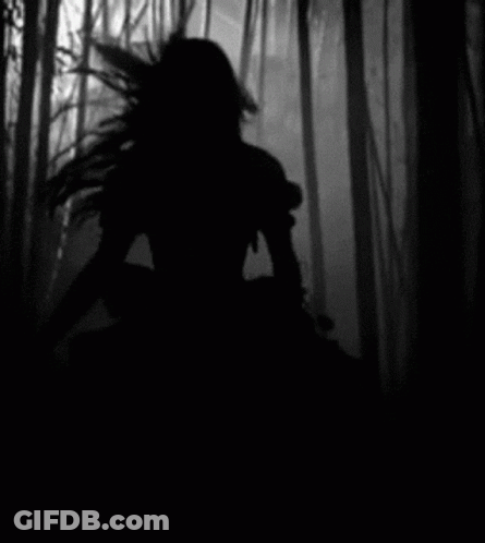 A Black And White Photo Of A Woman In A Dress Walking Through A Forest . GIF