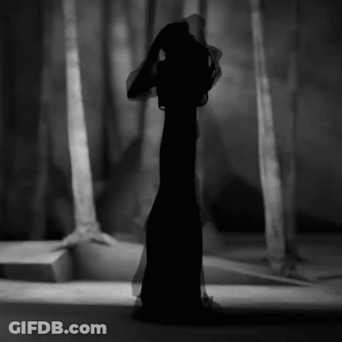 A Black And White Photo Of A Woman In A Long Black Dress In A Forest GIF