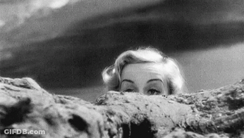 A Black And White Photo Of A Woman Peeking Over A Rock . GIF