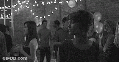 A Black And White Photo Of A Woman Standing In A Crowded Room At A Party . GIF