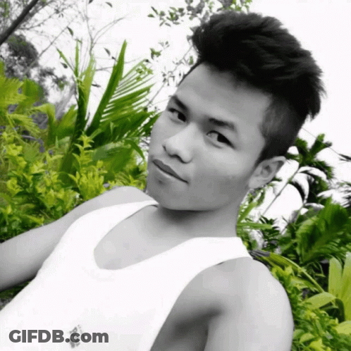 A Black And White Photo Of A Young Man Wearing A White Tank Top With A Few Plants Behind Him GIF
