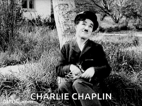 A Black And White Photo Of Charlie Chaplin Sitting Next To A Tree GIF