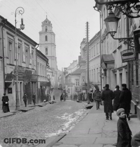 A Black And White Photo Of People Walking Down A Cobblestone Street With A Sign That Says ' Leningradska ' GIF