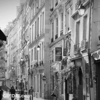 A Black And White Photo Of People Walking Down A Narrow Street With A Sign That Says Hotel Voyageur On It GIF