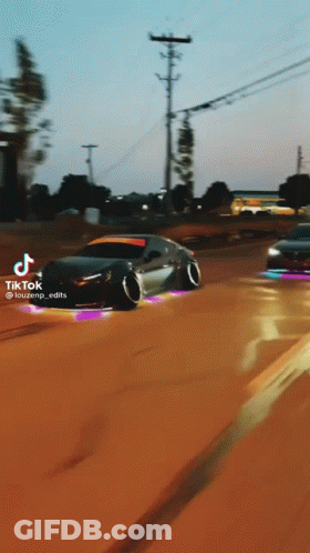 A Black Car Is Driving Down A Dirt Road With A School Bus In The Background .. GIF