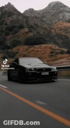 A Black Car Is Driving Down A Road In Front Of Mountains . GIF
