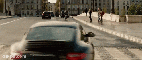 A Black Car Is Driving Down A Street With People Walking On The Sidewalk GIF