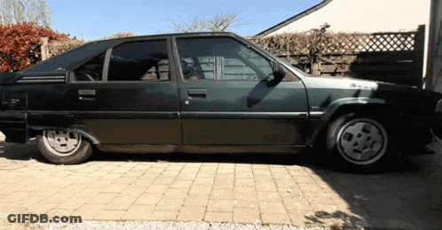 A Black Car Is Parked In A Driveway With A Fence In The Background GIF