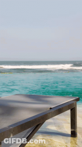 A Black Lounge Chair Sits On A Beach With The Ocean In The Background GIF