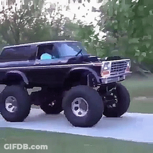 A Black Truck With Huge Tires Is Driving Down A Road . GIF
