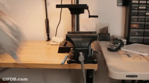 A Black Vise Sits On A Wooden Workbench Next To A Drill Press GIF