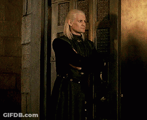 A Blonde Woman In A Black Coat Is Standing In Front Of A Door GIF