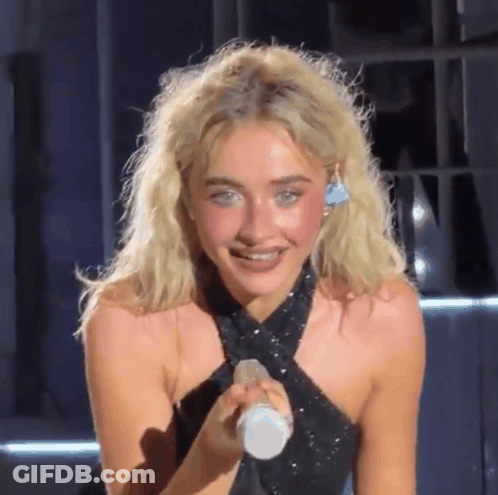 A Blonde Woman In A Black Dress Is Holding A Microphone And Smiling GIF
