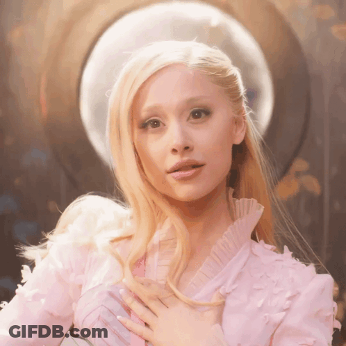 A Blonde Woman In A Pink Dress Holds Her Hands To Her Chest GIF