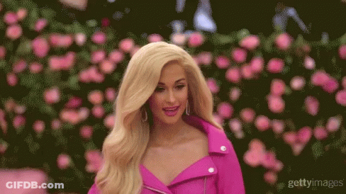 A Blonde Woman In A Pink Jacket Stands In Front Of A Wall Of Roses GIF