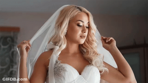 A Blonde Woman In A Wedding Dress Is Adjusting Her Veil GIF