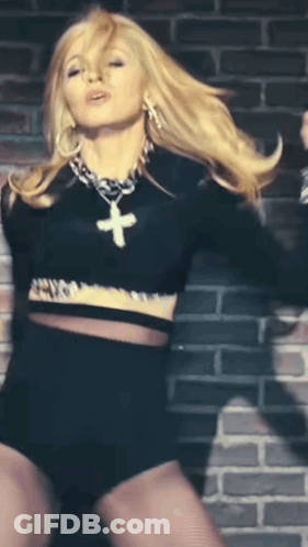 A Blonde Woman Wearing A Black Crop Top And Shorts With A Cross Necklace GIF