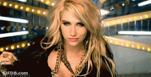 A Blonde Woman Wearing A Black Top And Gold Chains Looks At The Camera GIF