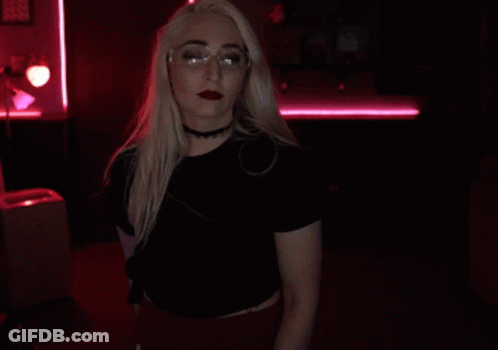 A Blonde Woman Wearing Glasses And A Choker Holds Her Finger To Her Lips GIF