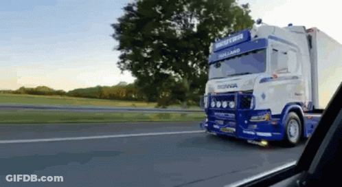 A Blue And White Truck Is Driving Down A Highway With Trees In The Background . GIF