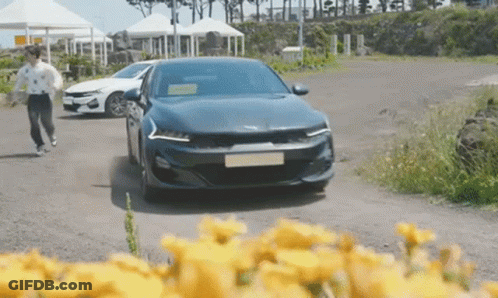 A Blue Car Is Driving Down A Dirt Road Next To A Field Of Yellow Flowers . GIF