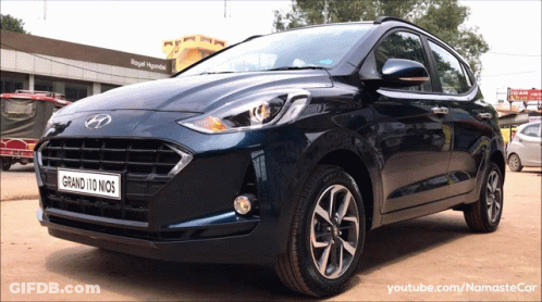 A Blue Hyundai Grand 110 Nios Is Parked In Front Of A Royal Hyundai Building GIF