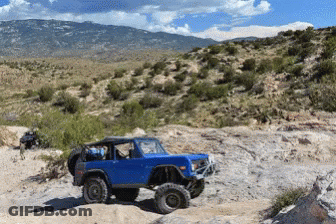 A Blue Jeep Is Parked On A Rocky Hillside With Mountains In The Background GIF