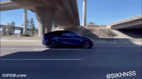 A Blue Tesla Model Y Is Driving Under A Highway Bridge GIF