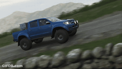 A Blue Truck Driving Down A Dirt Road With Mountains In The Background GIF