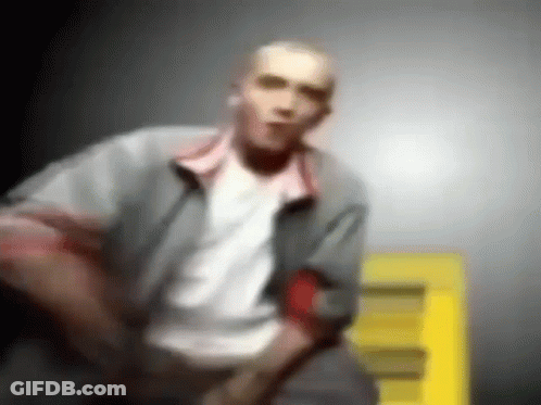 A Blurry Picture Of A Man In A Gray Jacket And White Shirt Standing In Front Of A Yellow Sign . GIF