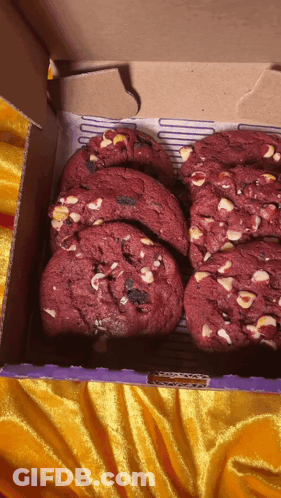 A Box Of Red Velvet Cookies With White Chocolate Chips Sits On A Yellow Cloth GIF
