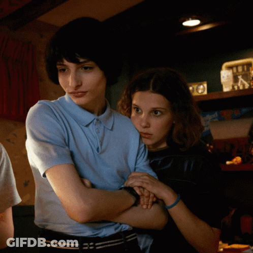 A Boy And A Girl Are Standing Next To Each Other With Their Arms Crossed In A Dark Room GIF