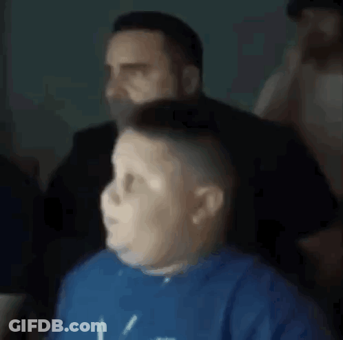 A Boy In A Blue Shirt Is Sitting In Front Of A Man In A Black Suit . GIF