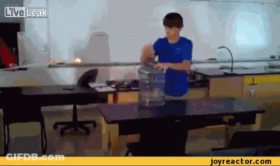A Boy In A Blue Shirt Is Standing In Front Of A Table With A Bucket Of Water On It .. GIF