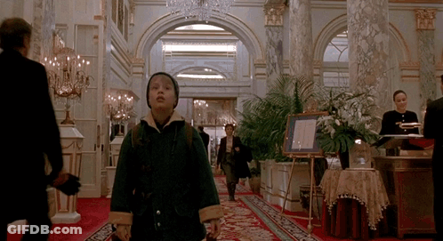 A Boy In A Green Coat Is Walking Down A Hallway In A Hotel GIF