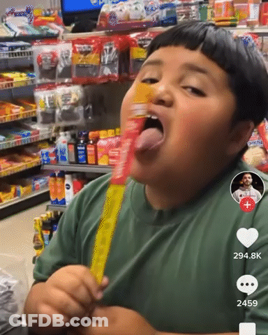A Boy In A Green Shirt Is Eating A Yellow Candy Bar In A Store GIF