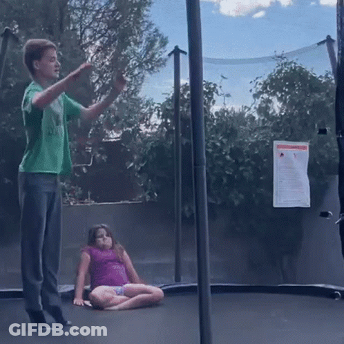 A Boy In A Green Shirt Is Jumping On A Trampoline While A Girl In A Pink Shirt Sits On The Ground . GIF