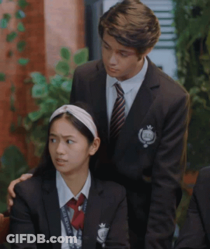A Boy In A Suit And Tie Stands Next To A Girl In A School Uniform With A Lanyard That Says Homemaker On It GIF