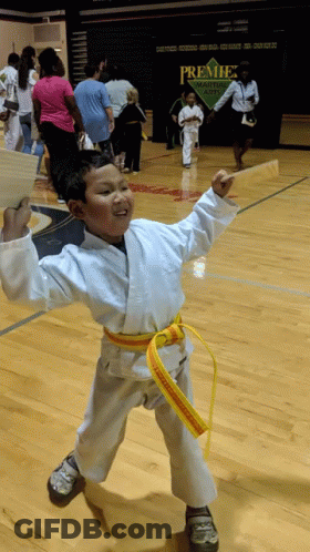 A Boy In A White Karate Uniform With A Yellow Belt Stands In Front Of A Sign That Says Premier Martial Arts GIF
