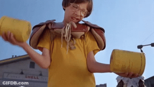 A Boy In A Yellow Shirt Is Holding Two Yellow Cans In Front Of A Shell Store GIF