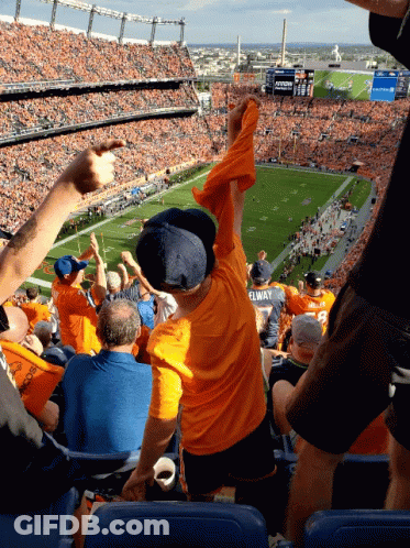A Boy In A Yellow Shirt With Elway On The Back Of It Stands In The Stands At A Football Game GIF