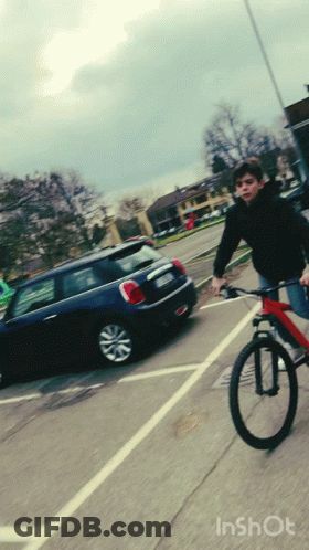 A Boy Is Riding A Bike In A Parking Lot With A Mini Cooper Behind Him GIF